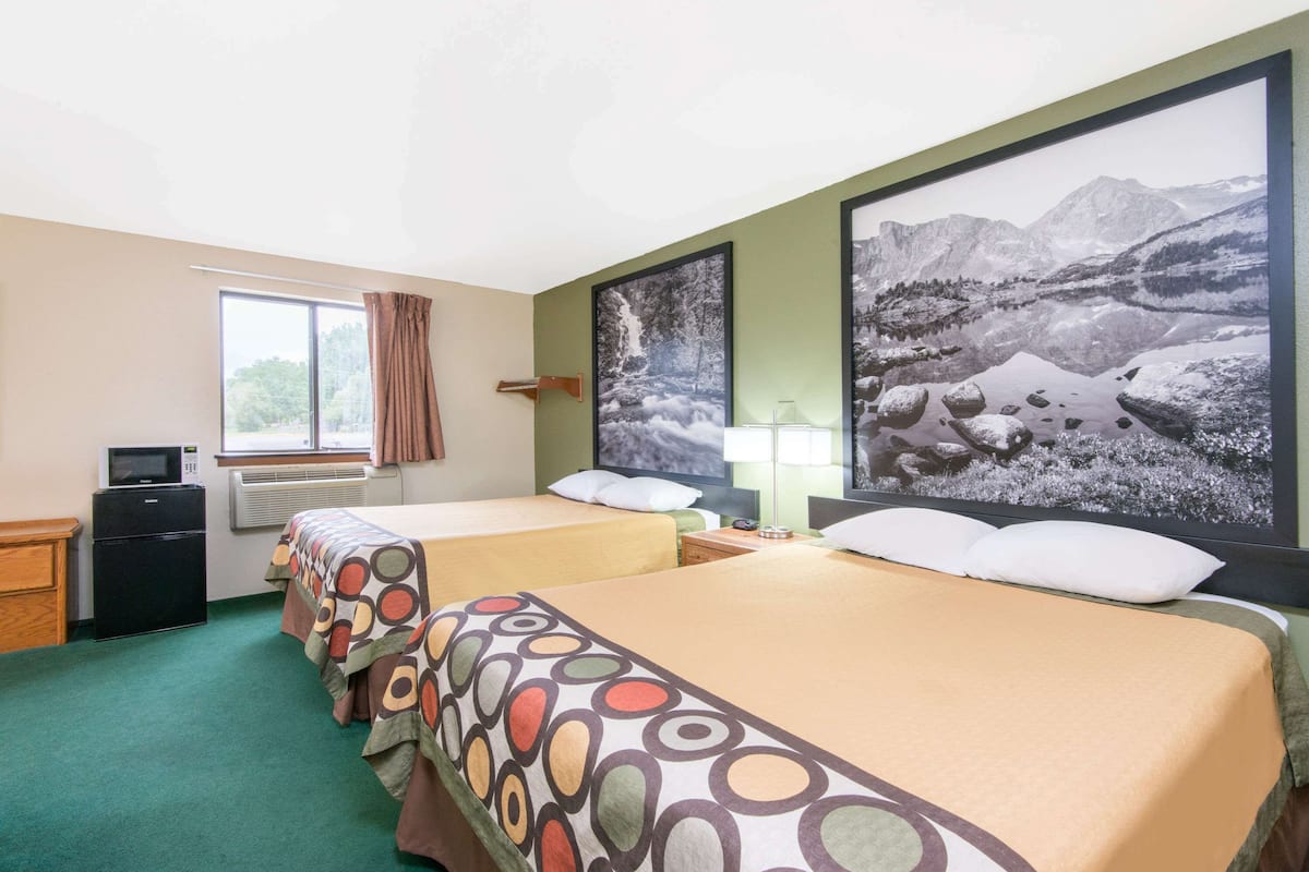 Ein Hotelzimmer mit zwei Betten, einem Kühlschrank, einer Mikrowelle, einem Fenster mit Vorhängen und einem großen gerahmten Bild einer Berglandschaft.