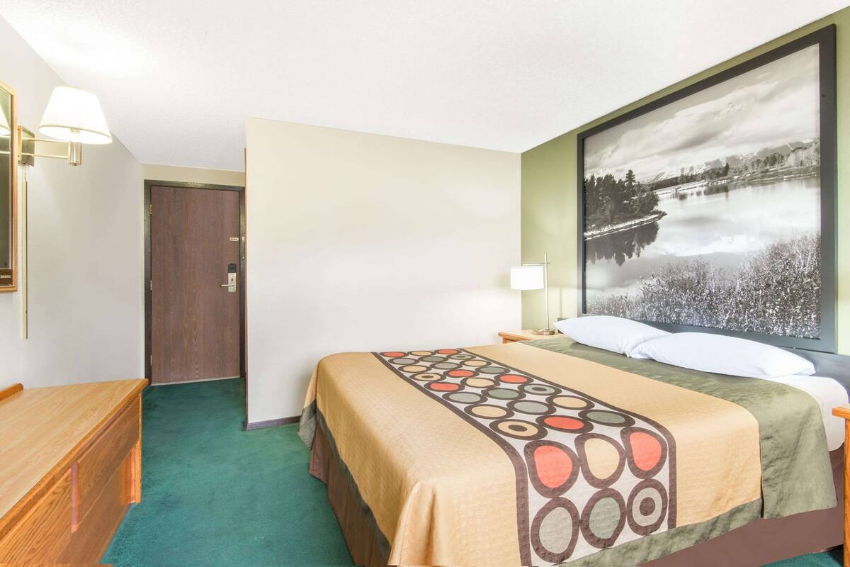 Ein Hotelzimmer mit Bett, Schreibtisch, Stuhl, Lampe und einem großen, gerahmten Bild eines Sees.