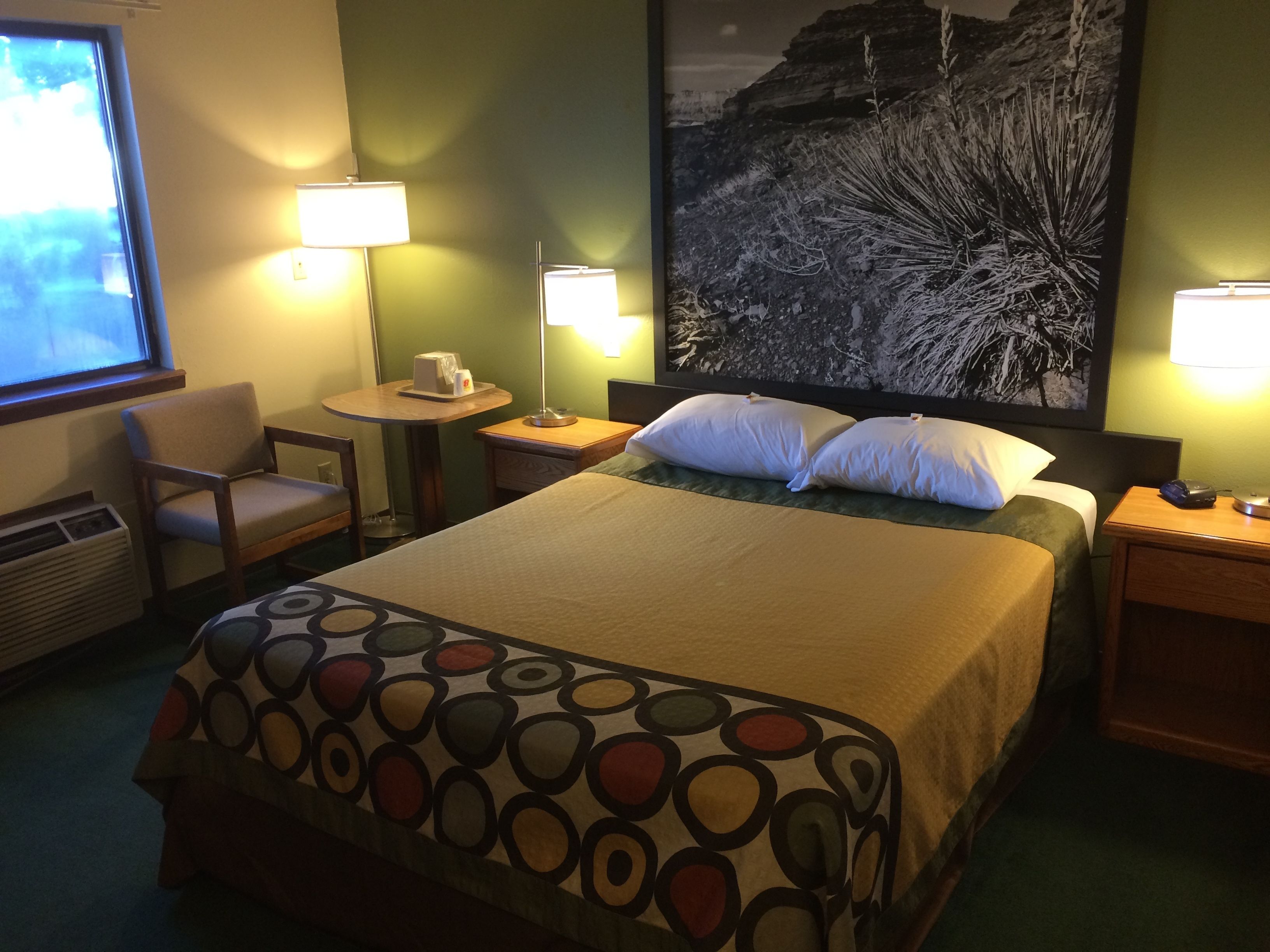 Quarto de hotel com cama, duas luminárias de cabeceira, uma mesinha com cafeteira, uma cadeira e um quadro grande na parede.