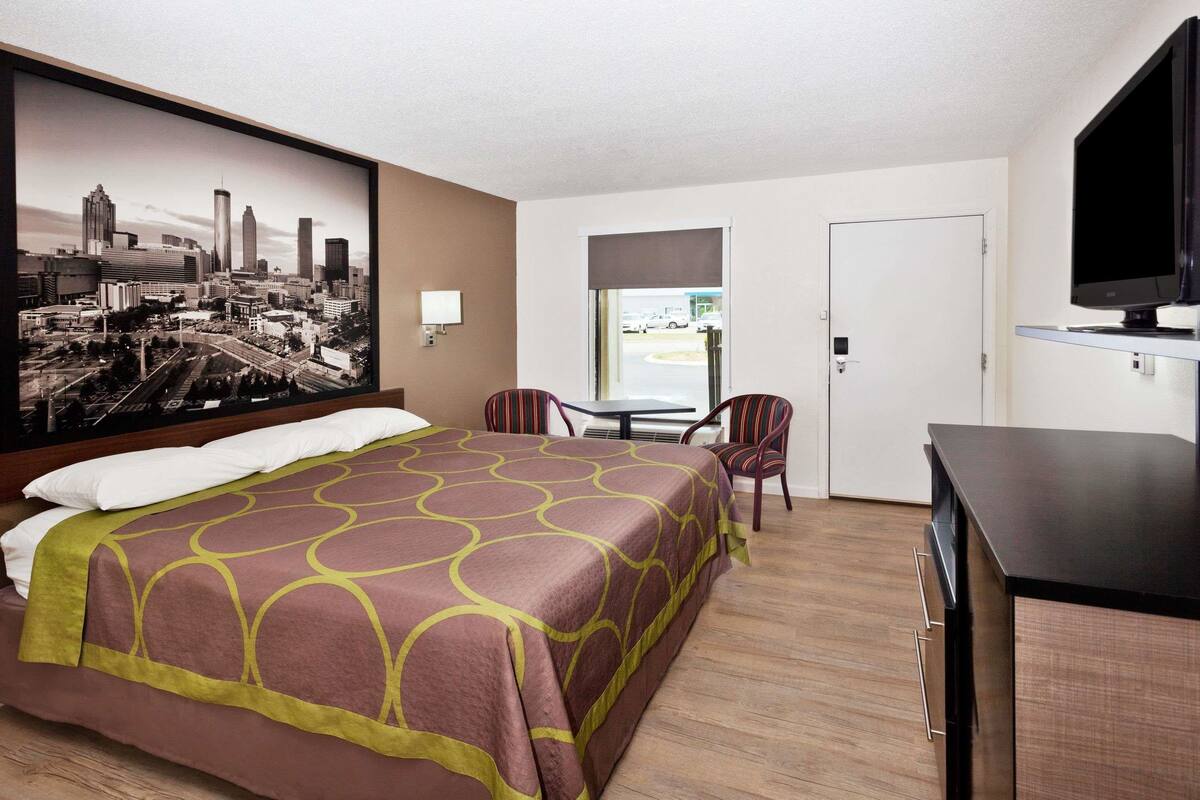 Une chambre d’hôtel avec un lit, un bureau, une télévision, une chaise et un tableau représentant un paysage urbain accroché au mur.