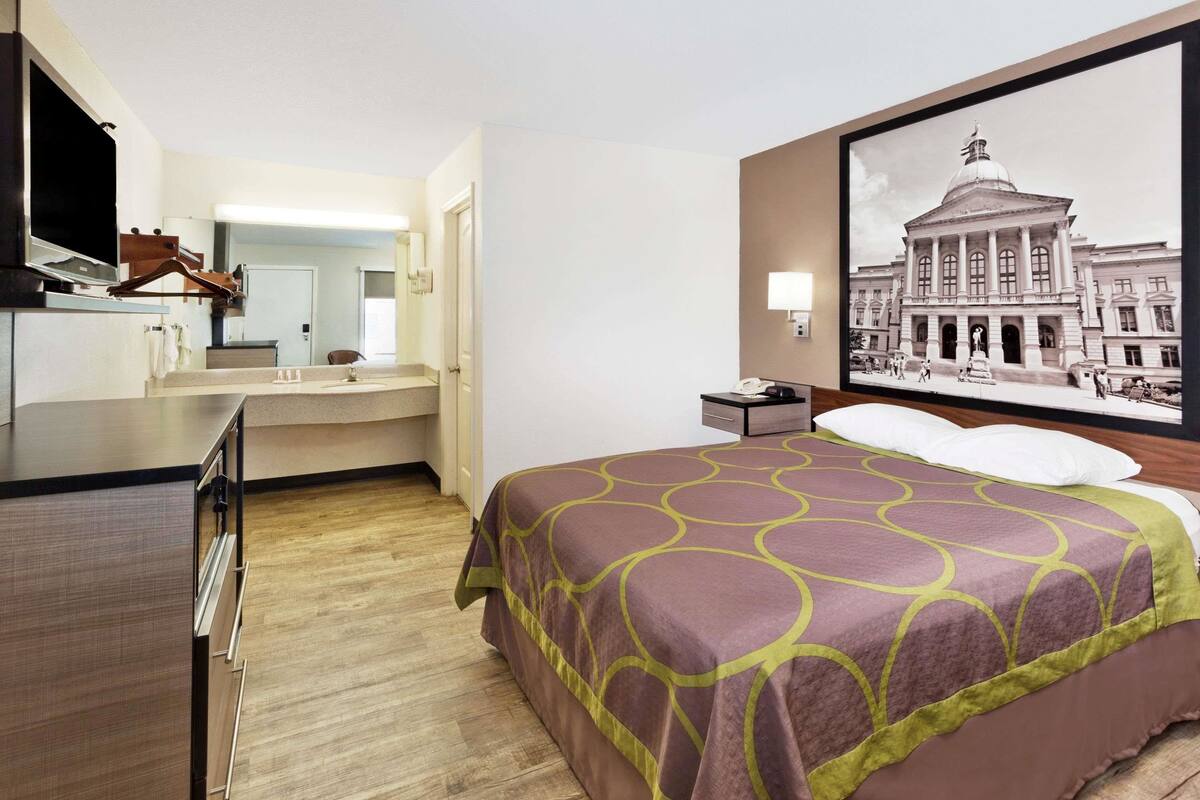 Une chambre d’hôtel avec un lit, un bureau, un miroir, une télévision et un grand tableau encadré au mur.