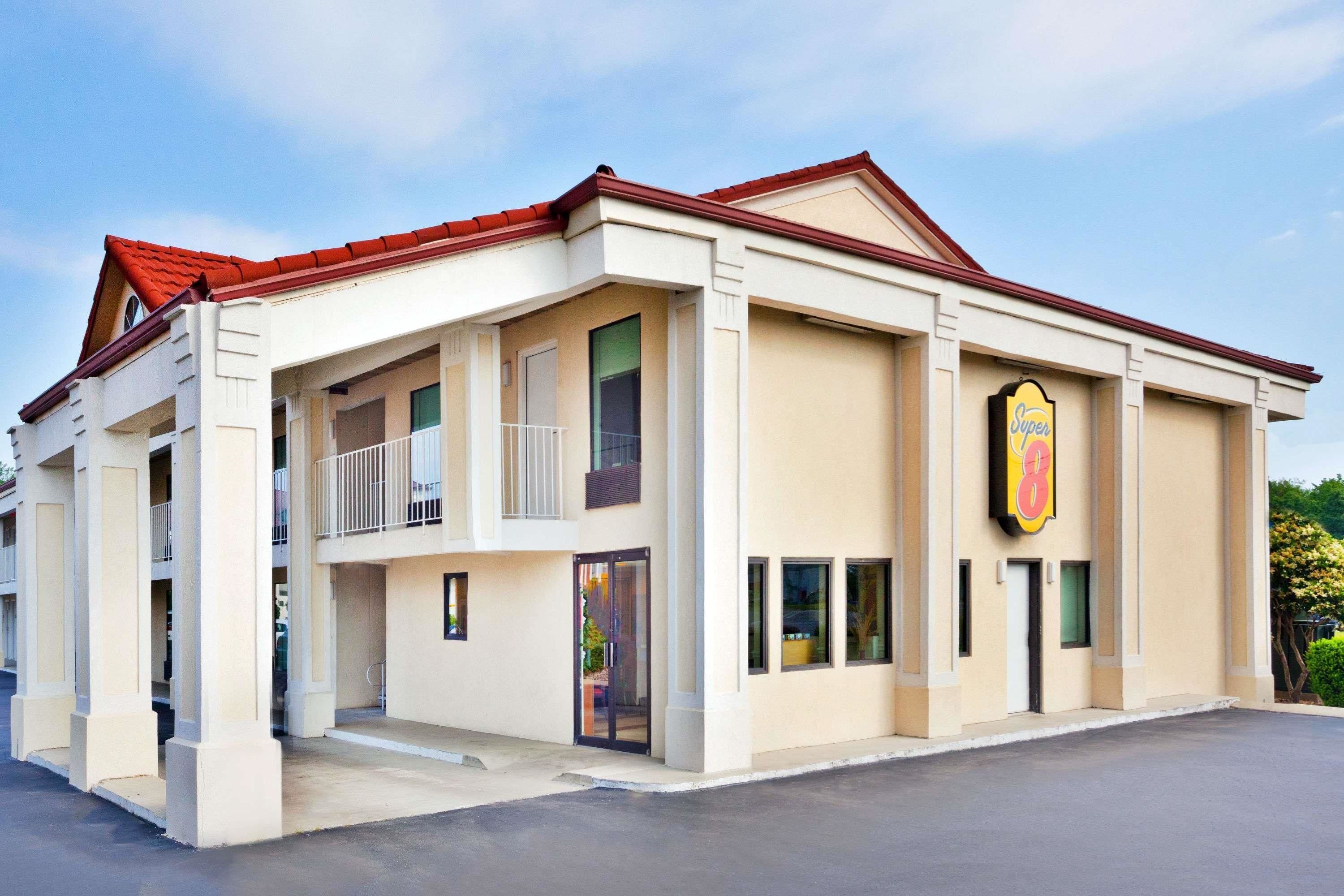A hotel with a beige facade, a red roof, and a prominent 'Super 8' sign. It features a balcony on the second floor and a covered entrance.