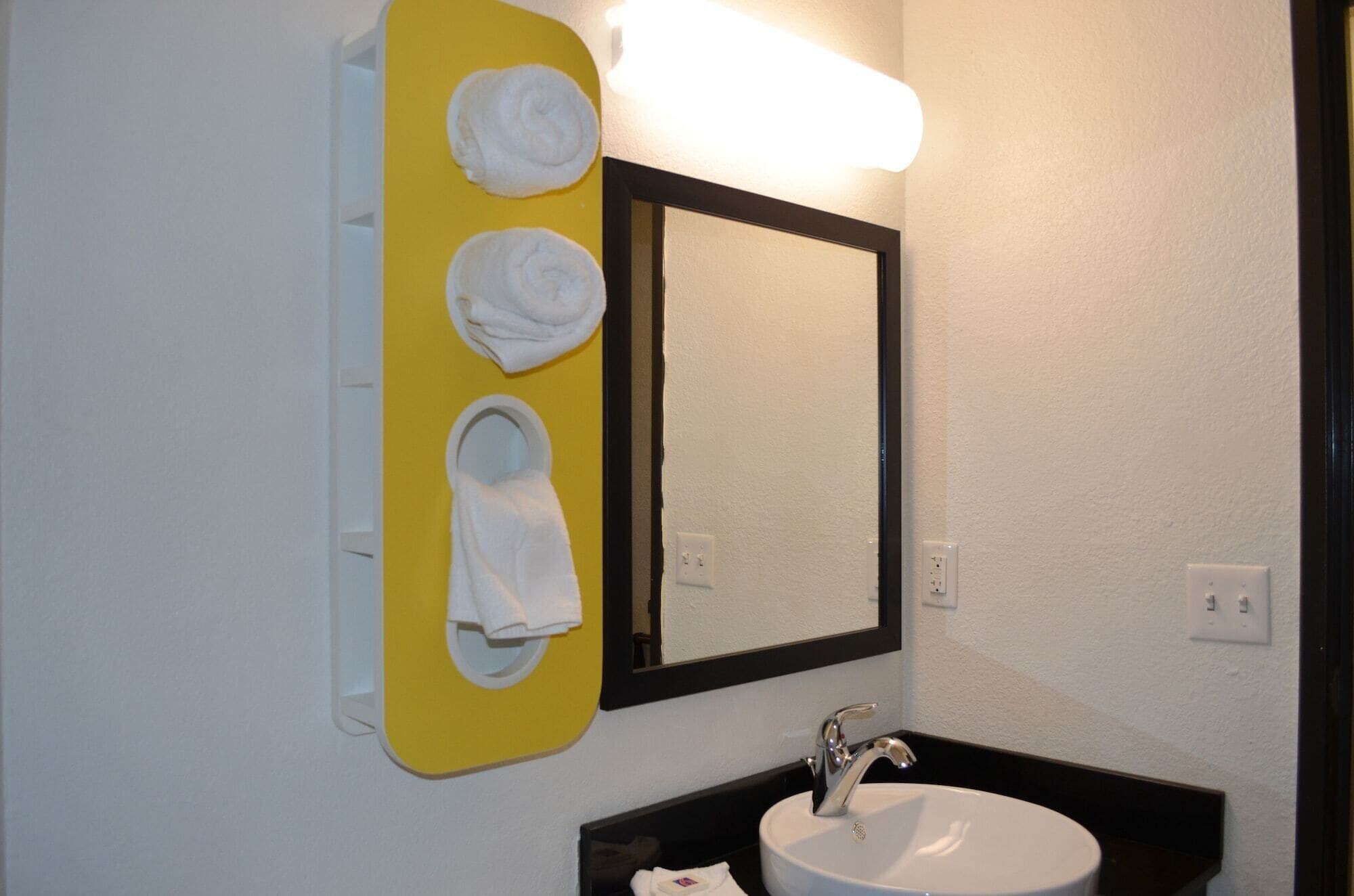 A bathroom with a yellow towel rack holding three white towels, a white sink with a chrome faucet, and a mirror with a black frame.