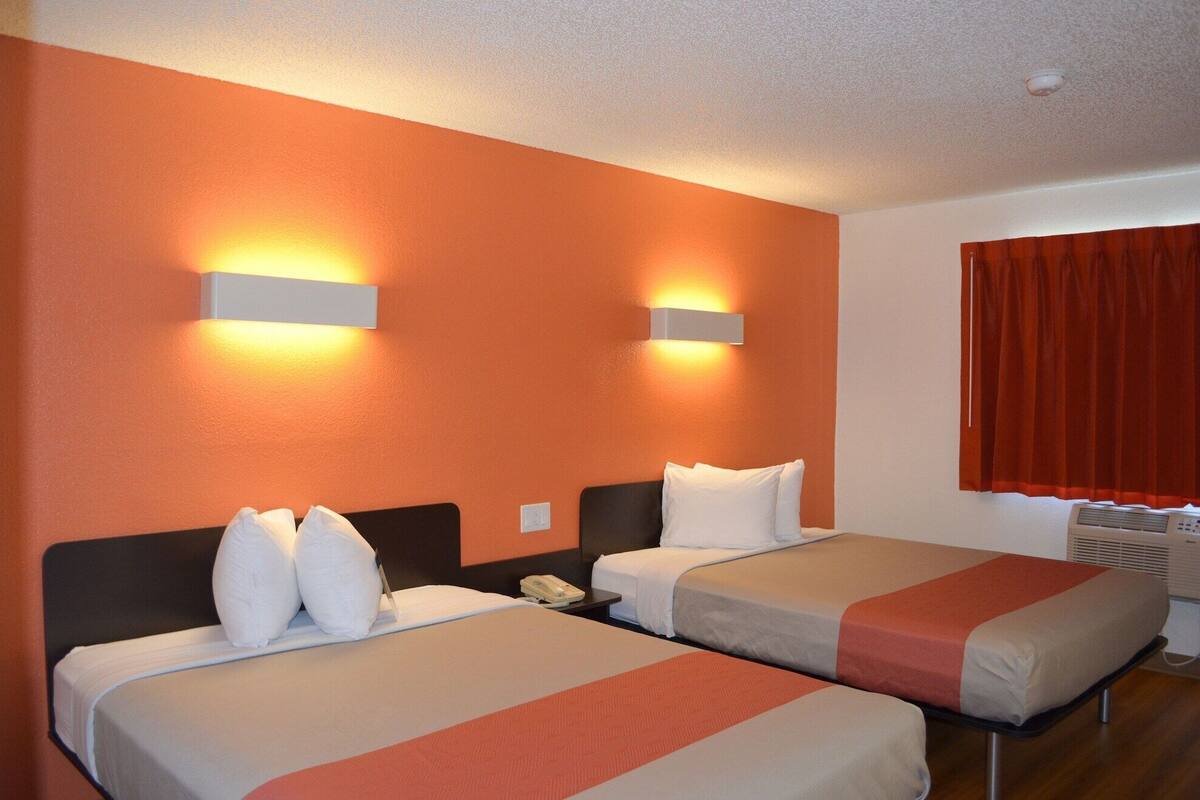 A hotel room with two beds, each with white bedding and a red accent. There are wall-mounted light fixtures on the orange walls.