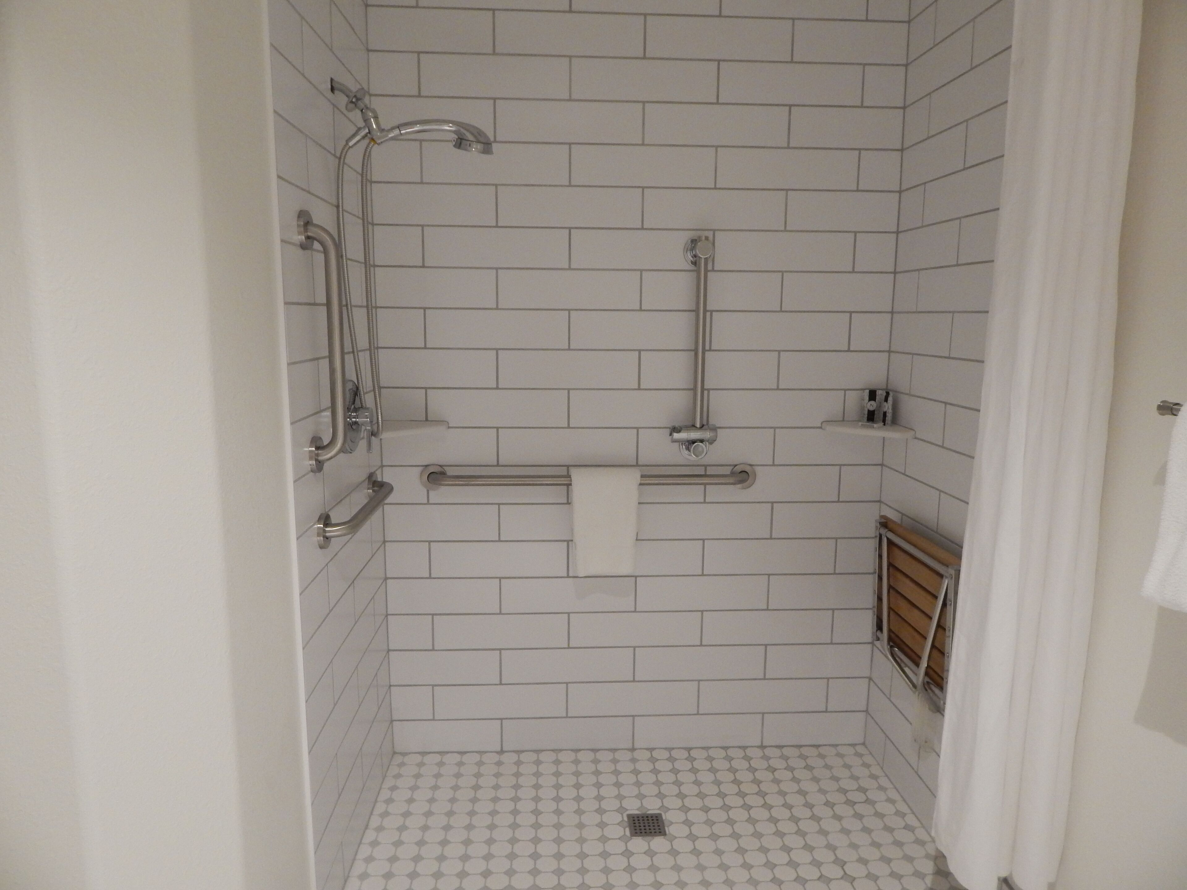 Un baño pequeño con azulejos blancos, ducha de mano, barra de apoyo y toallero.