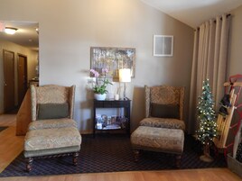 Una sala de estar con dos sillones estampados, una mesita con una lámpara y una planta, una estantería, un árbol de Navidad y una escalera roja.