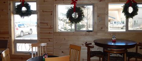 A wooden cabin interior with a dining area, two windows with wreaths, and a view of the outside.