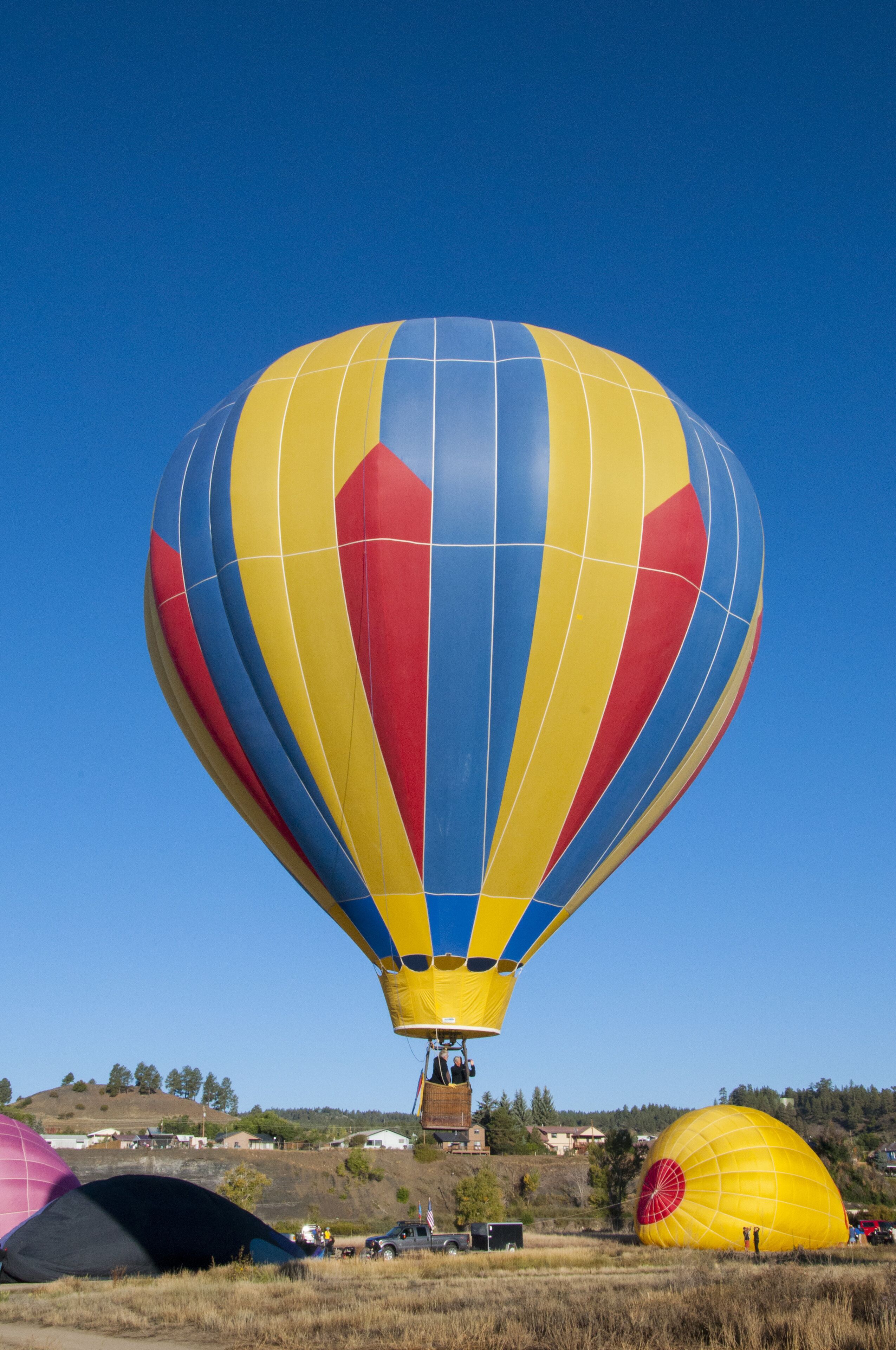 A hot air balloon with a yellow, blue, and red design, anchored in a field with other balloons in the background.
