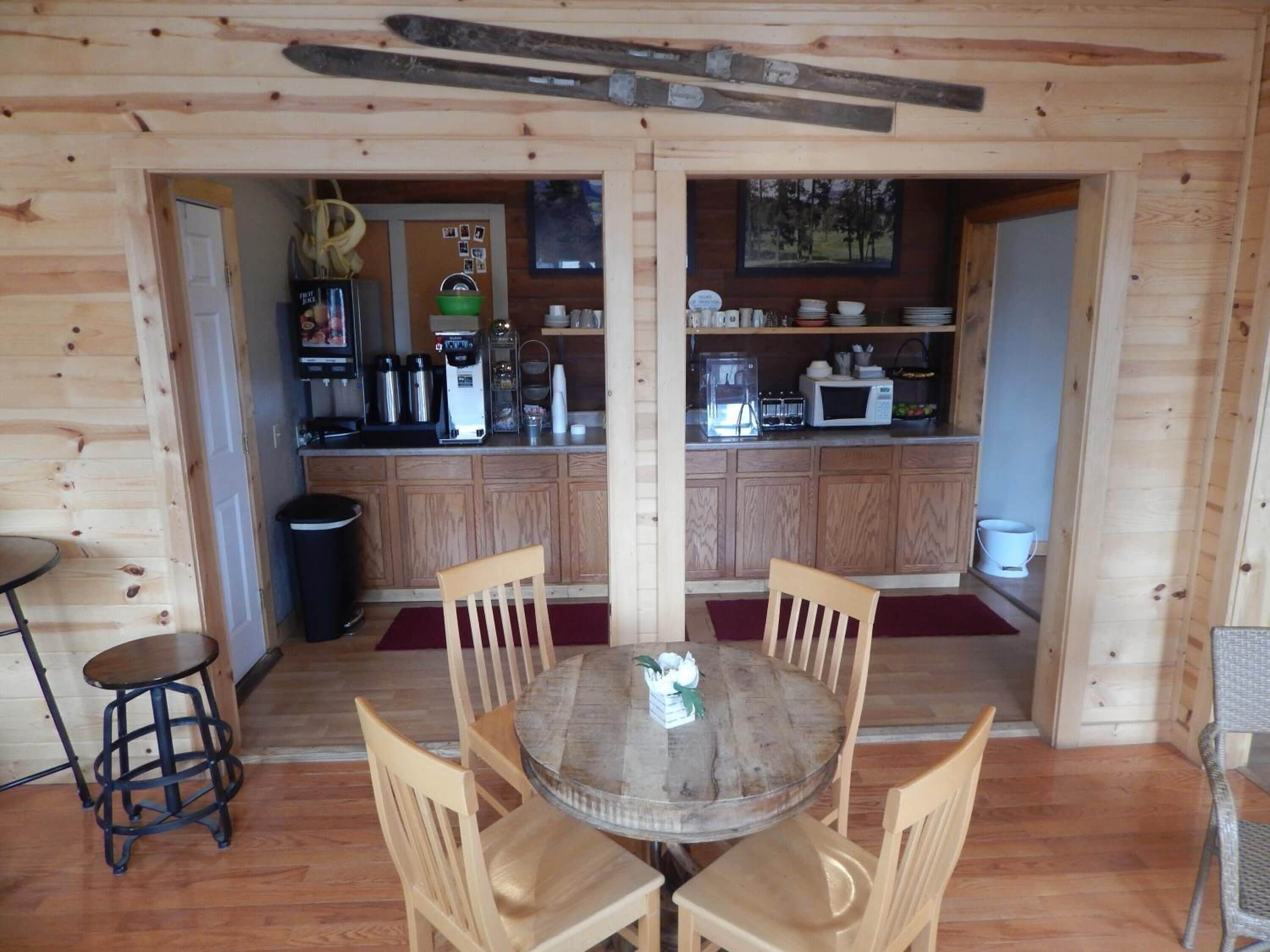 Een houten blokhut interieur met een ronde eettafel, vier stoelen, een keuken met apparatuur, en een ski-rek aan het plafond.