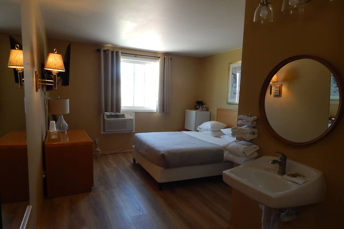 A hotel room with a bed, a bedside table with a lamp, a small desk, a mirror, and a bathroom with a sink and toilet.