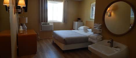Ett hotellrum med en säng, ett sängbord med en lampa, ett litet skrivbord, en spegel och ett badrum med handfat och toalett.