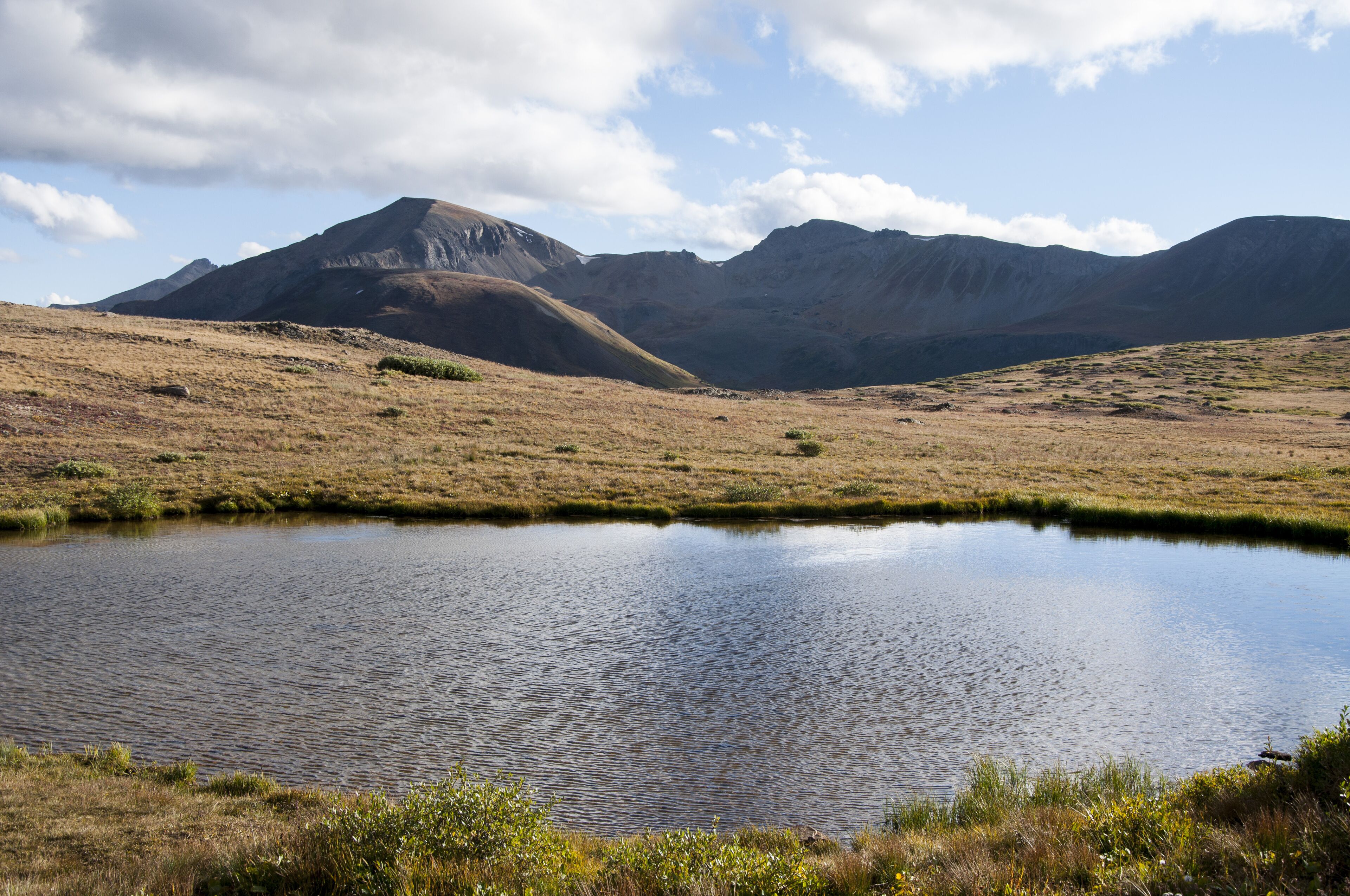 Un paisaje sereno con un pequeño estanque, colinas cubiertas de hierba y montañas lejanas bajo un cielo parcialmente nublado.