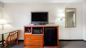 Habitación de hotel con televisión, microondas, refrigerador, mueble de madera, una silla y una mesa con lámpara.