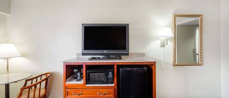 A hotel room with a television, microwave, refrigerator, wooden cabinet, chair, and a table with a lamp.