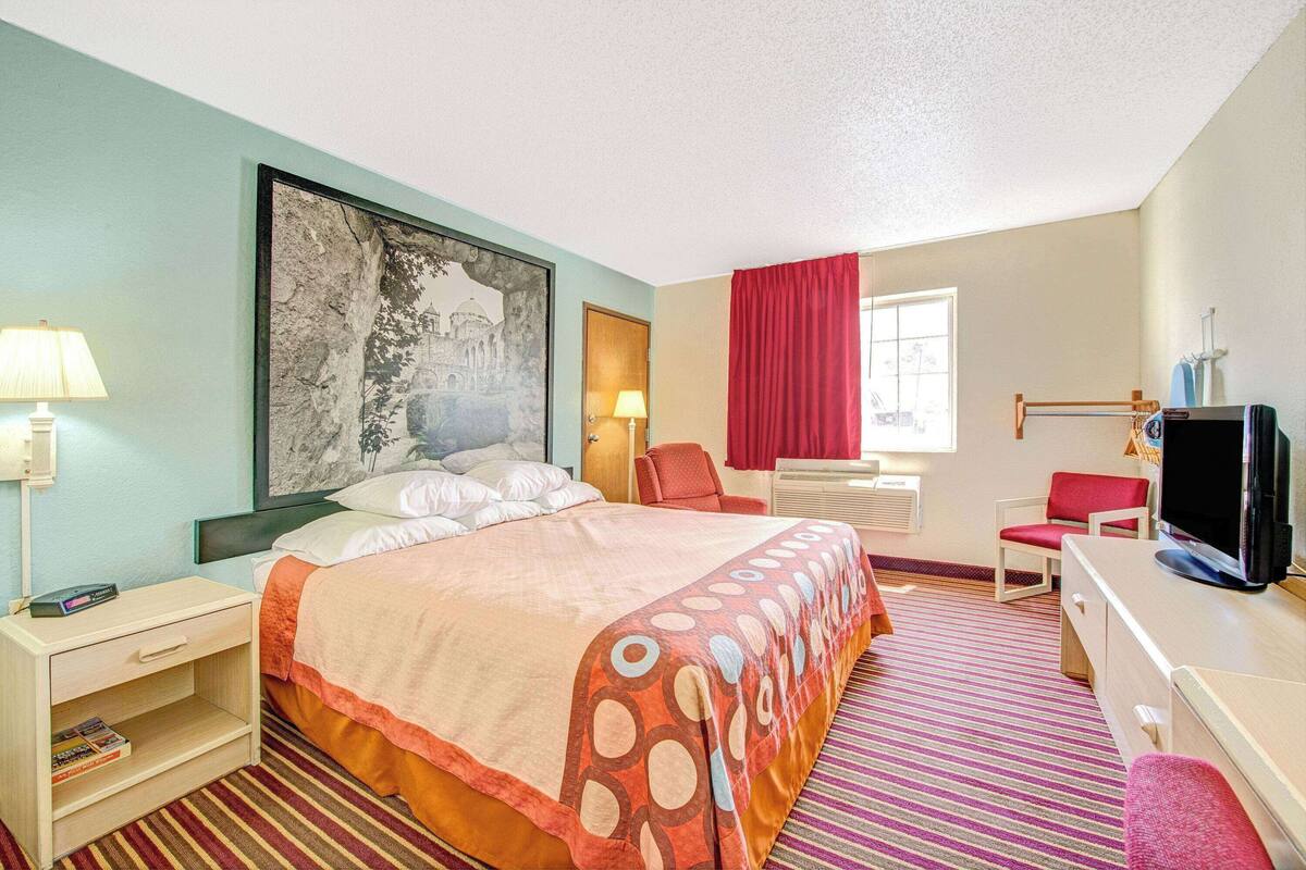 Ein Hotelzimmer mit Bett, Schreibtisch mit Fernseher, Sessel, Lampe und einem großen Gemälde an der Wand.