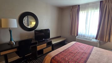 A hotel room with a bed, a desk with a computer, a television, a mirror, a window with curtains, and an air conditioning unit.