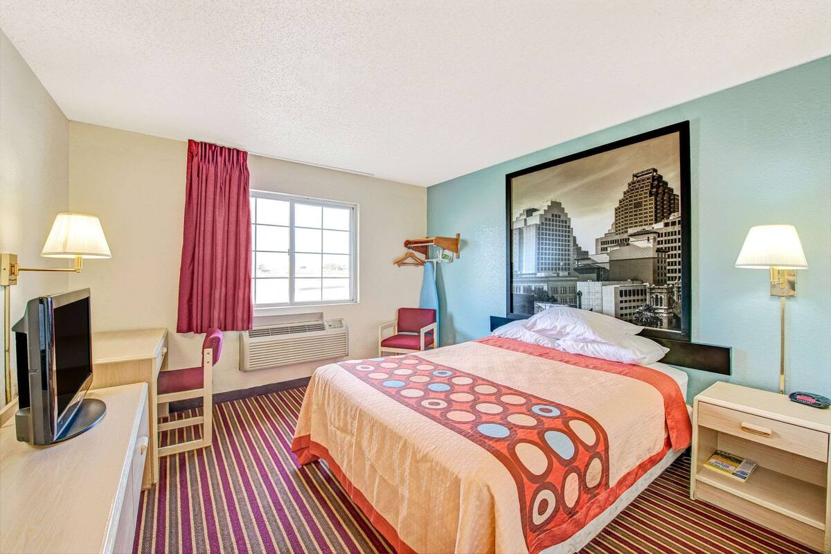 Ein Hotelzimmer mit Bett, Schreibtisch, Stuhl, Fernseher, Lampe und einem fenster mit Vorhängen.