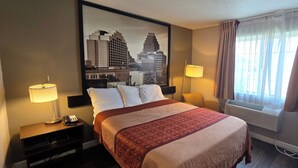 A hotel room with a large bed, two bedside lamps, a cityscape painting on the wall, an air conditioning unit, and a window with curtains. - Super 8 by Wyndham San Antonio/Fiesta (San Antonio)