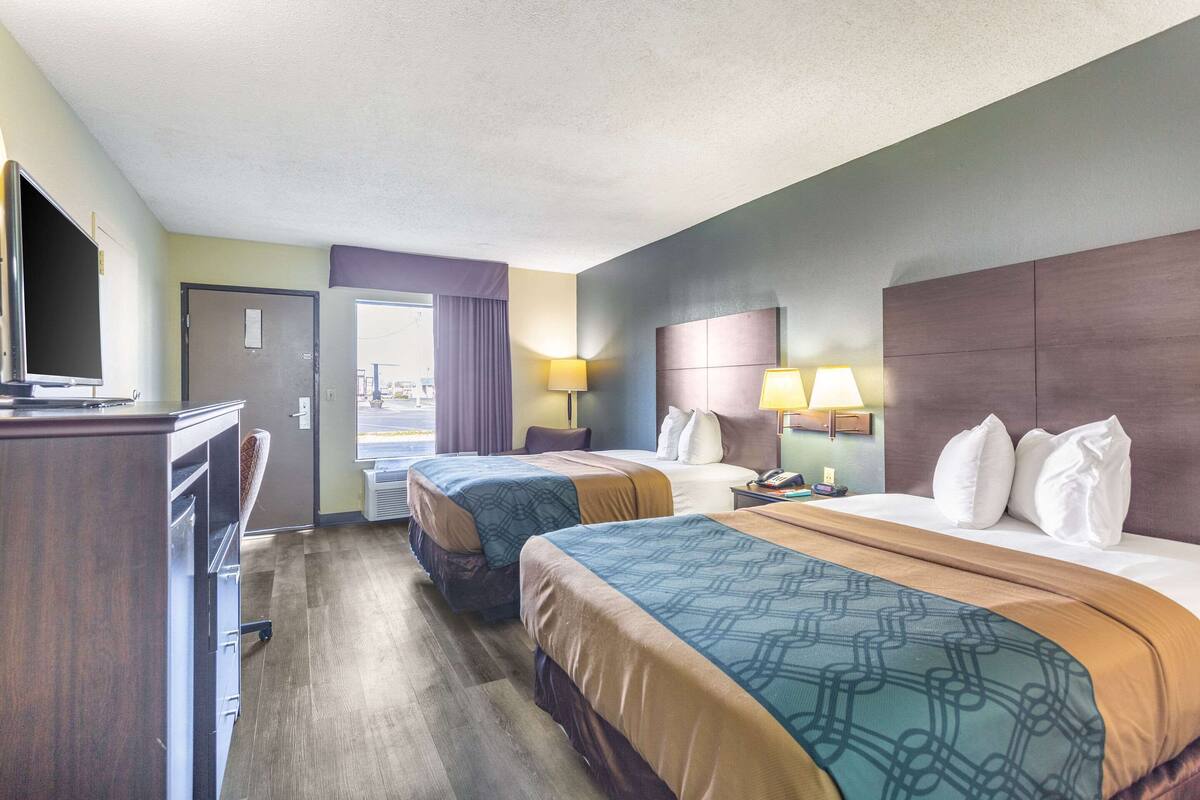 A hotel room with two beds, a desk with a flat-screen TV, and a window with curtains.