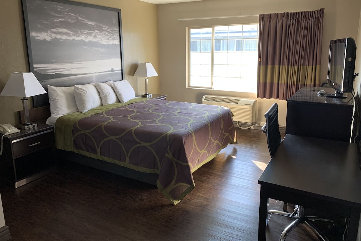 A hotel room with a bed, two bedside lamps, a desk with a computer, a window with curtains, and a wall-mounted air conditioner.
