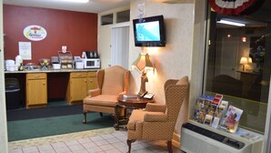 A hotel room with two patterned armchairs, a small table with a lamp, a television mounted on the wall, and a window with magazines on the windowsill.