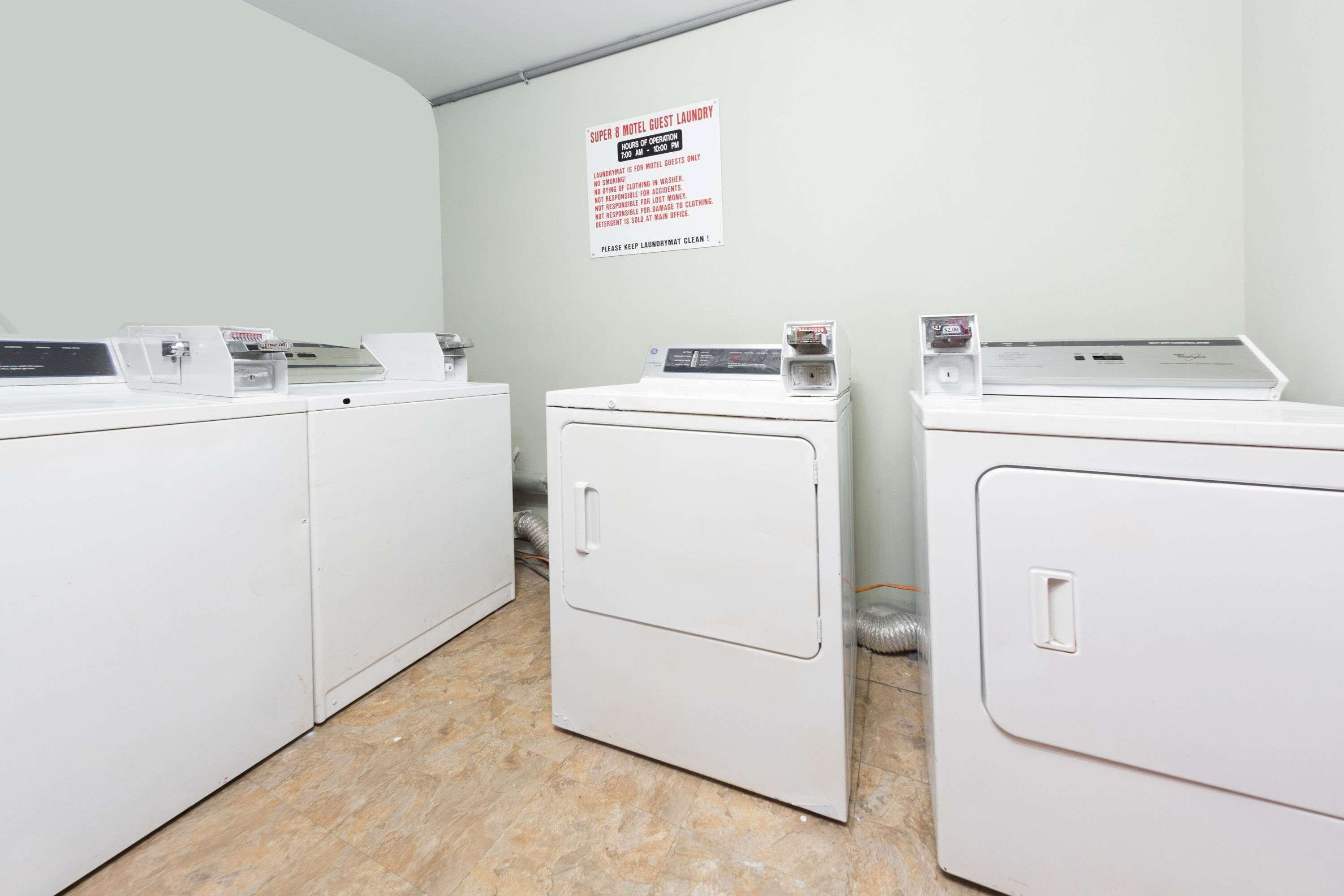 A laundry area with three white washing machines, a sign indicating guest laundry hours, and a vent on the wall.