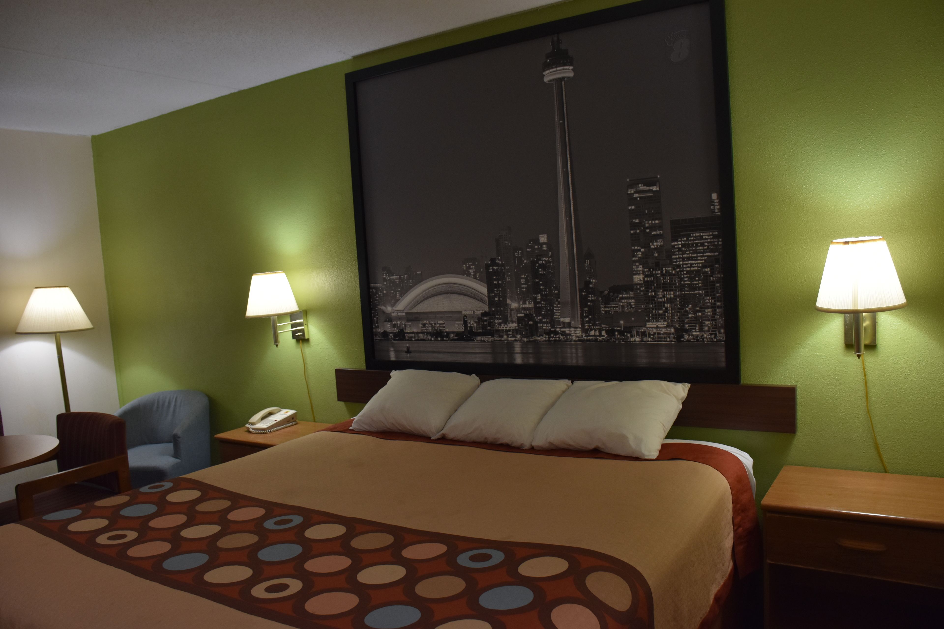 A hotel room with a bed, two bedside lamps, a chair, a desk, and a cityscape painting on the wall.