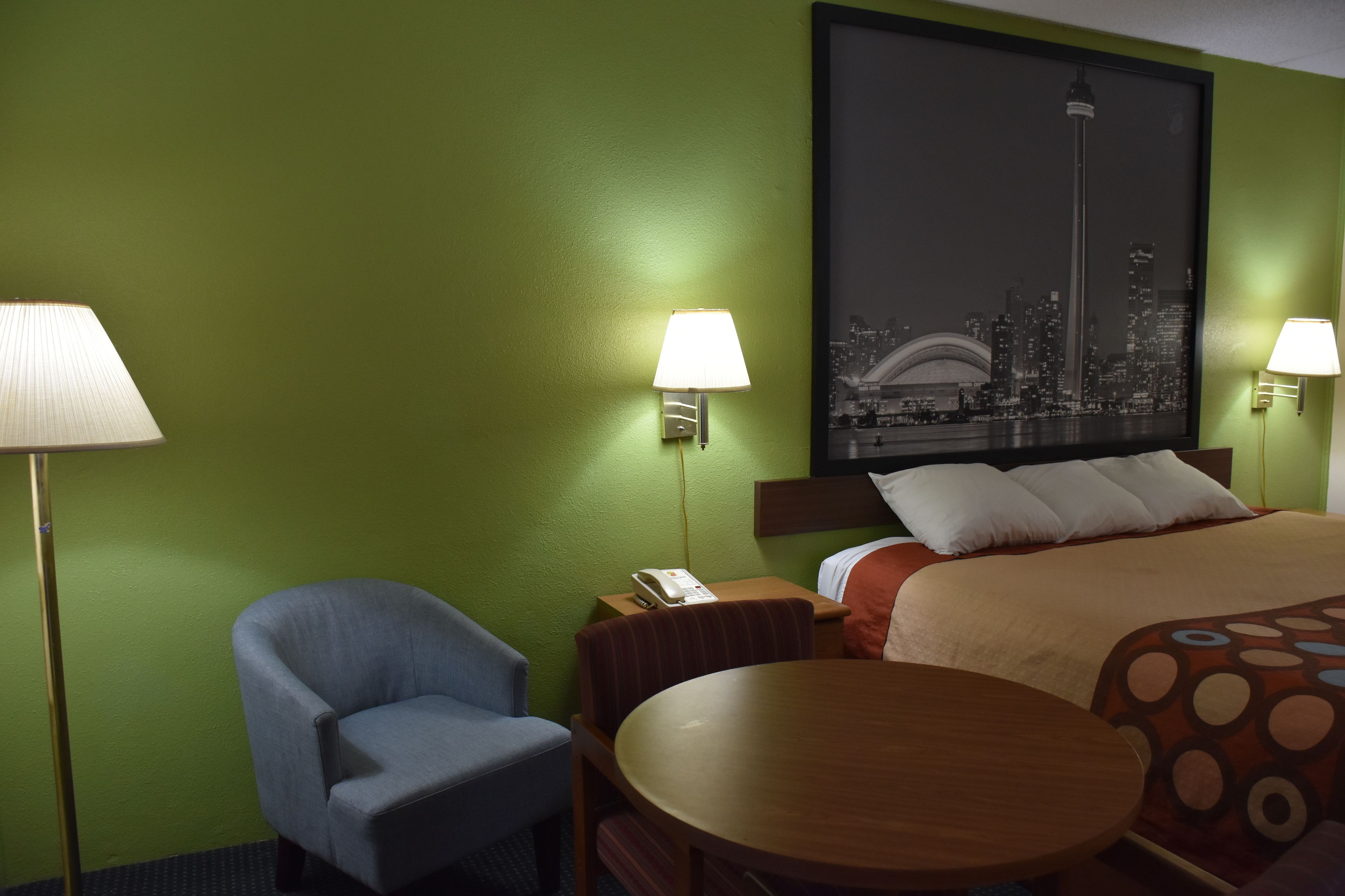 A hotel room with a green wall, a bed with a patterned blanket, a wooden table, a blue armchair, and two bedside lamps.