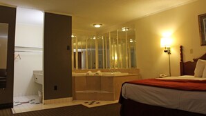 A hotel room with a bed, a large mirror, a bathroom with a bathtub, and a nightstand with a lamp.