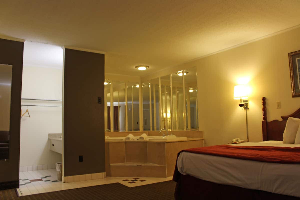 Een hotelkamer met een bed, een grote spiegel, een badkamer met een bad en een nachtkastje met een lamp.