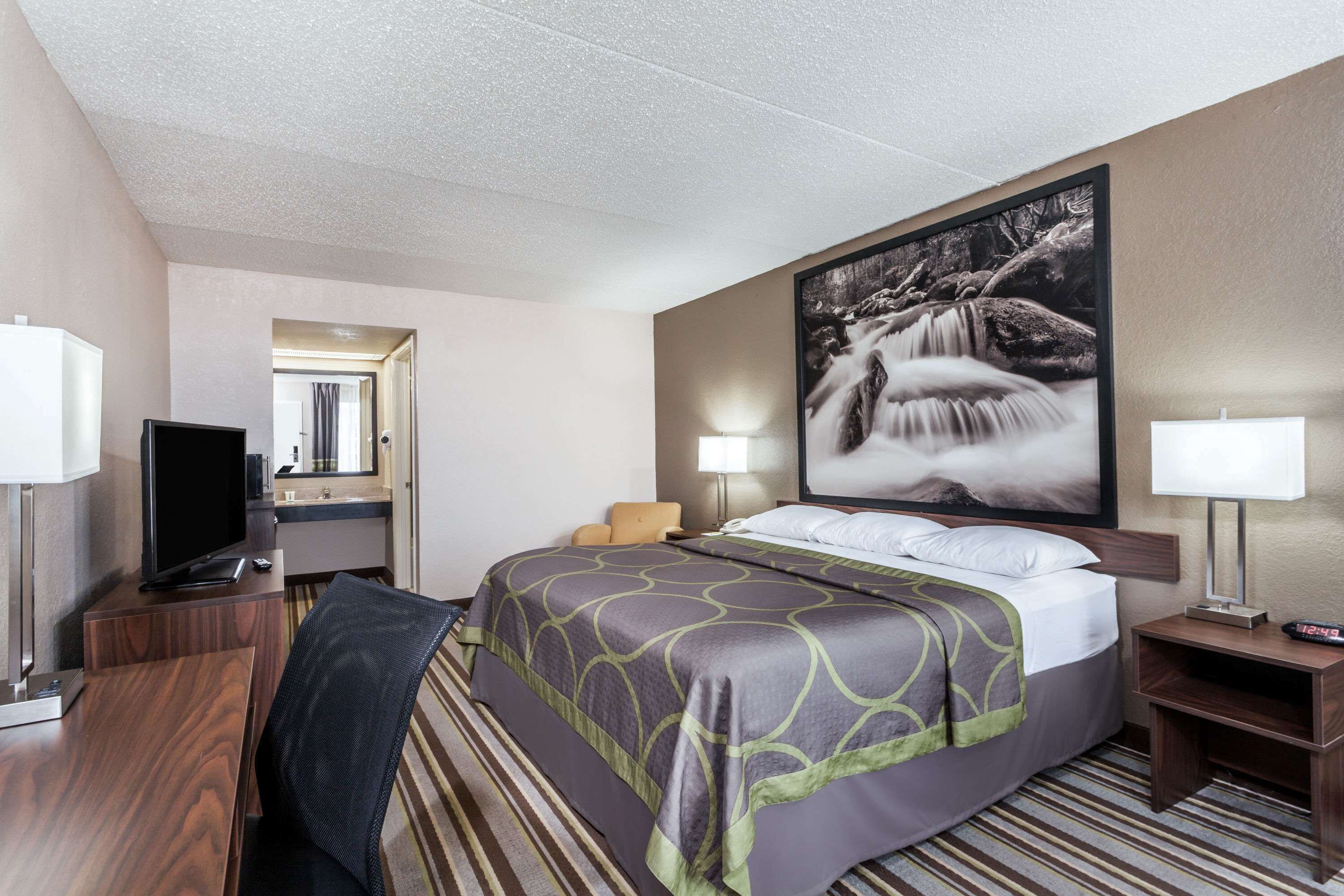 A hotel room with a bed, a desk with a computer, a TV, a bathroom mirror, and a large framed picture on the wall.