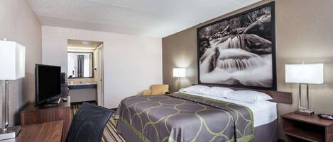 A hotel room with a bed, a desk with a computer, a TV, a bathroom mirror, and a large framed picture on the wall.