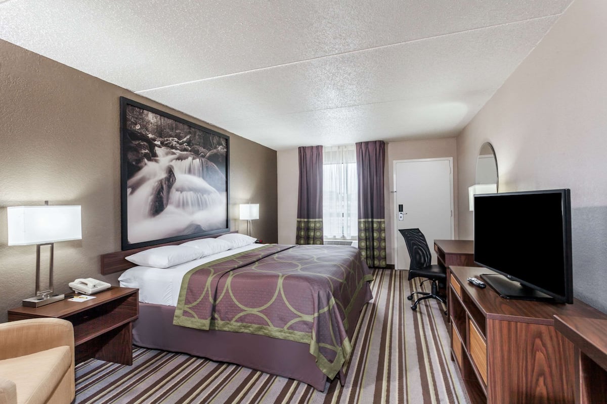A hotel room with a bed, a desk with a lamp, a television, and a large painting on the wall.