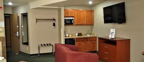 A hotel room with a bed, a red armchair, a desk, a flat-screen TV, wooden cabinets, a microwave, and a small kitchenette.