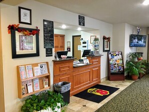 Zona de recepción de un hotel con mostrador de madera, un cubo de basura, certificados enmarcados, un panel de menú y un felpudo con la marca 'Super8'.