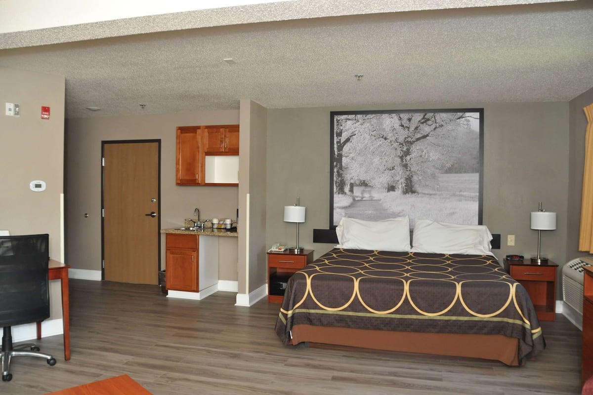 A hotel room with a bed, a desk, a chair, a small kitchenette, and a large window with a view of trees.