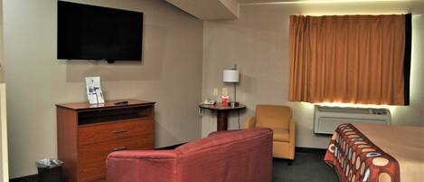 Habitación de hotel con televisor de pantalla plana en la pared, cómoda de madera, sofá rojo, mesita de noche con lámpara, una silla y cama con colcha estampada.