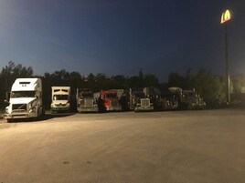 Uma fileira de caminhões estacionados num estacionamento à noite, com um letreiro do McDonald's ao fundo.