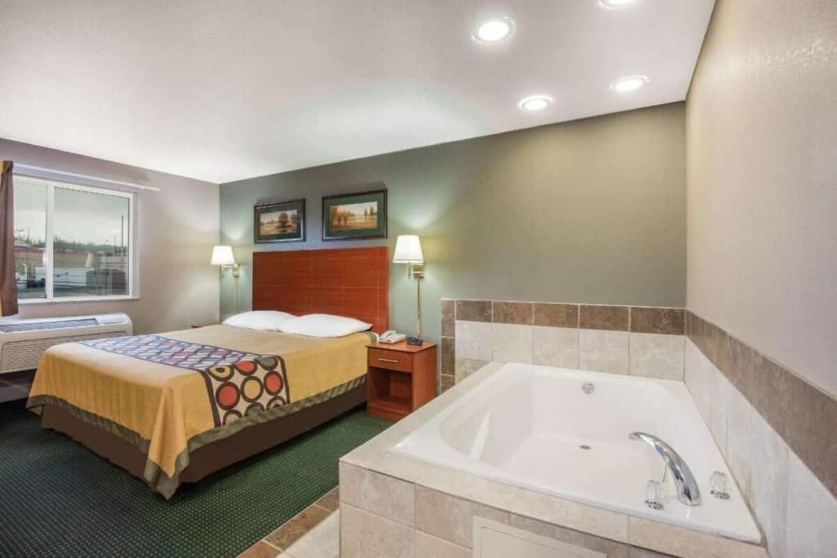 A hotel room with a bed, a window, a wall-mounted heater, a bedside table with a lamp, and a bathtub.