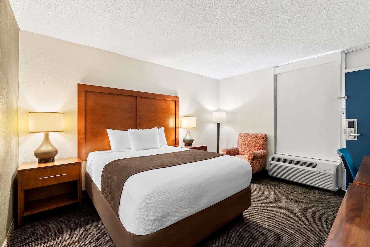 A hotel room with a large bed, two bedside tables with lamps, a chair, a desk, and an air conditioning unit.