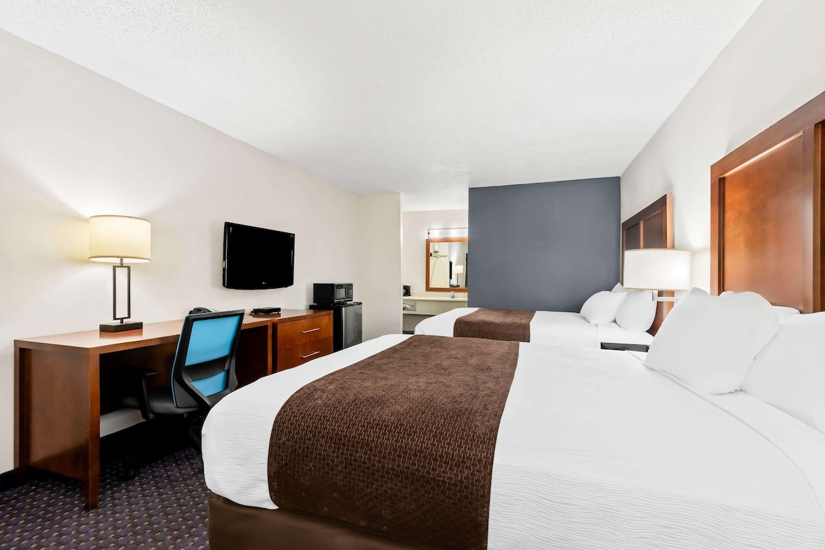 A hotel room with two beds, a desk with a chair, a television, and a bathroom visible in the background.