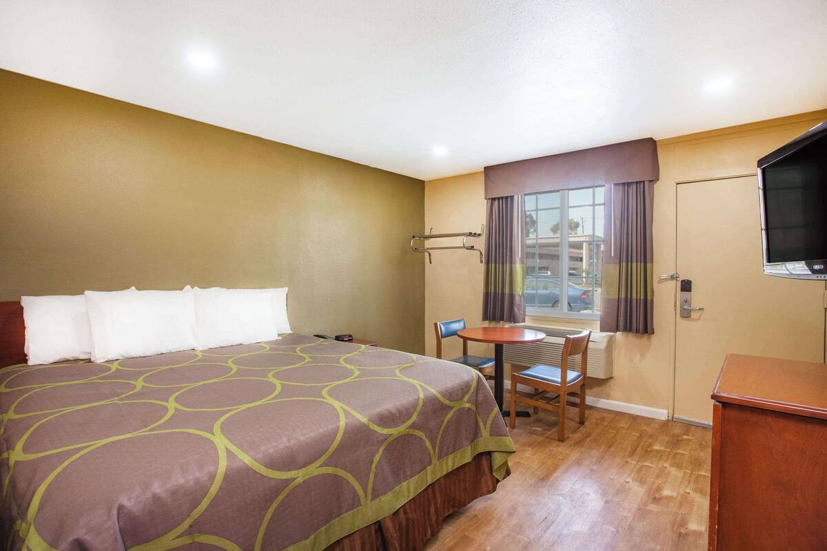 A hotel room with a large bed, a small table with a chair, a TV, and a window with curtains.