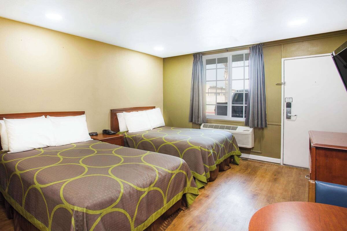 A hotel room with two beds, a wooden floor, a window with curtains, a door, and a small table.