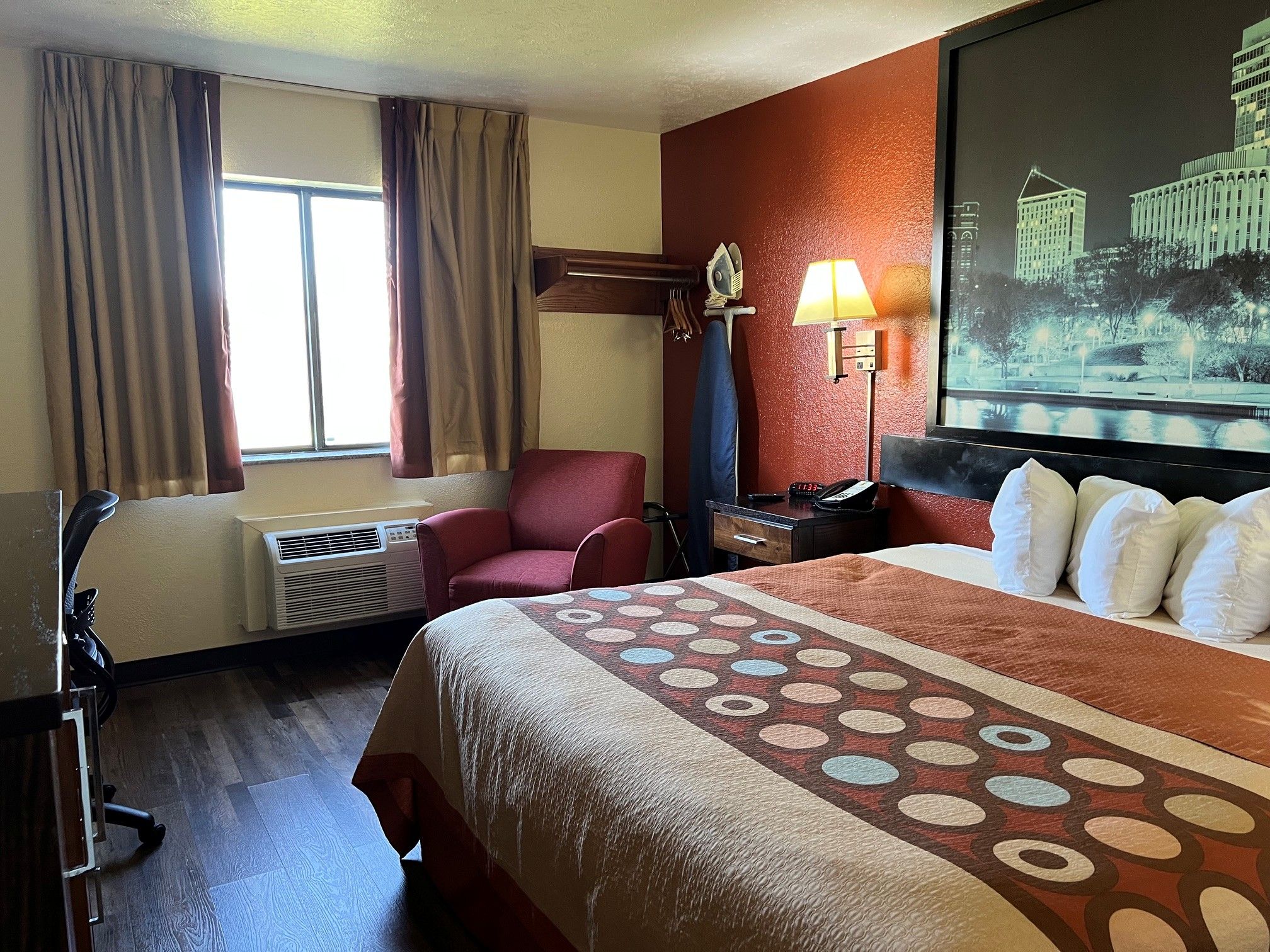 Een hotelkamer met een bed, een bureau, een stoel, een raam met gordijnen, een airconditioning, een lamp en een schilderij aan de muur.