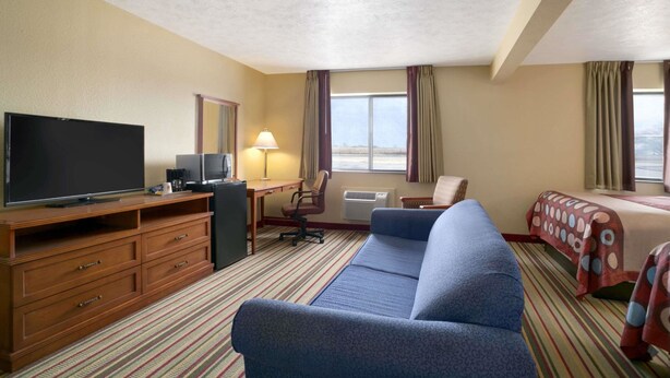 Quarto de hotel com uma televisão de tela plana grande, cômoda de madeira, sofá azul, escrivaninha com luminária e uma cama com colcha listrada.
