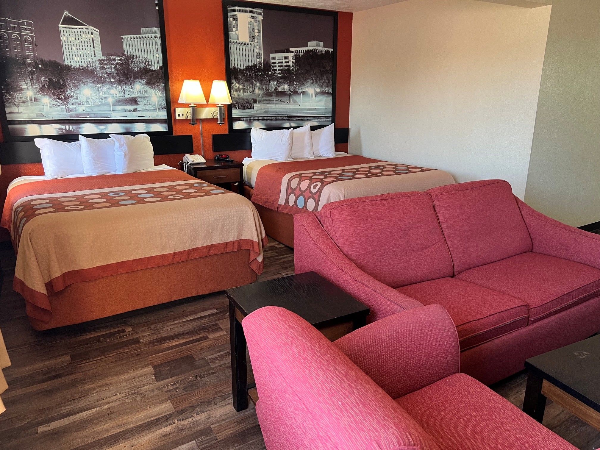 A hotel room with two beds, a red sofa, a small table, and a nightstand with a lamp.