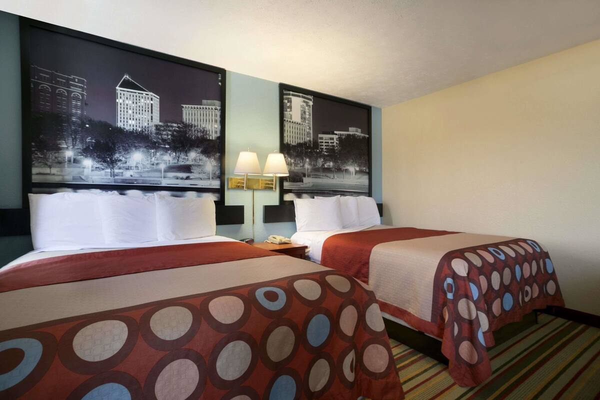 Habitación de hotel con dos camas, una mesita de noche con lámpara y un cuadro de un paisaje urbano en la pared.