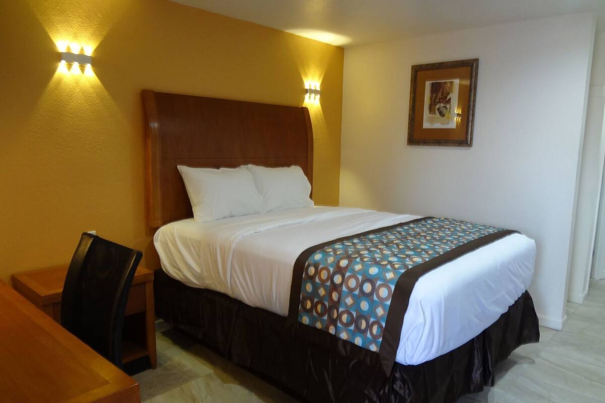 A hotel room with a wooden headboard, a bed with white linens, a patterned throw blanket, and a desk with a chair.