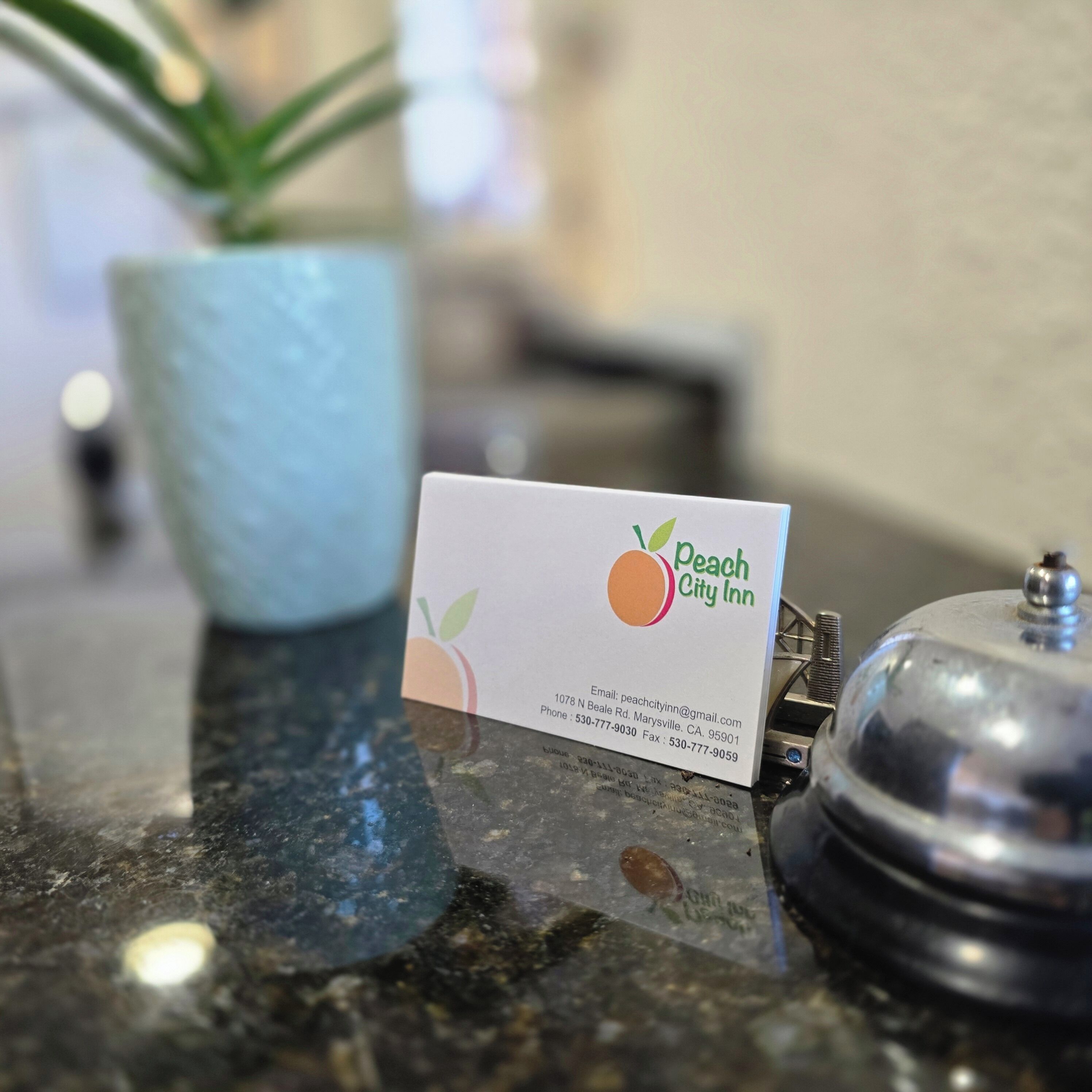 Une carte de visite du Peach City Inn, posée sur un comptoir en marbre, avec une plante en pot et une cloche en métal en arrière-plan.