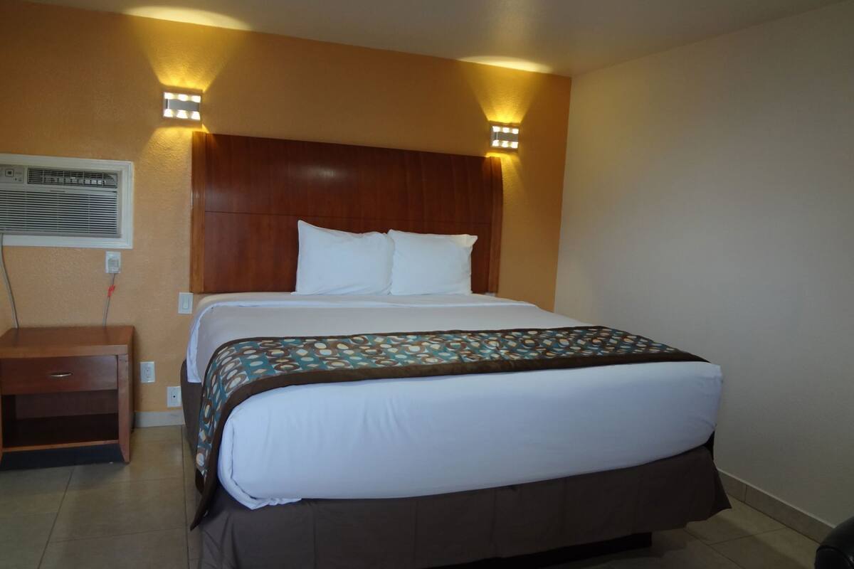 A hotel room with a large bed, a wooden headboard, two wall-mounted lights, a bedside table, and an air conditioning unit.